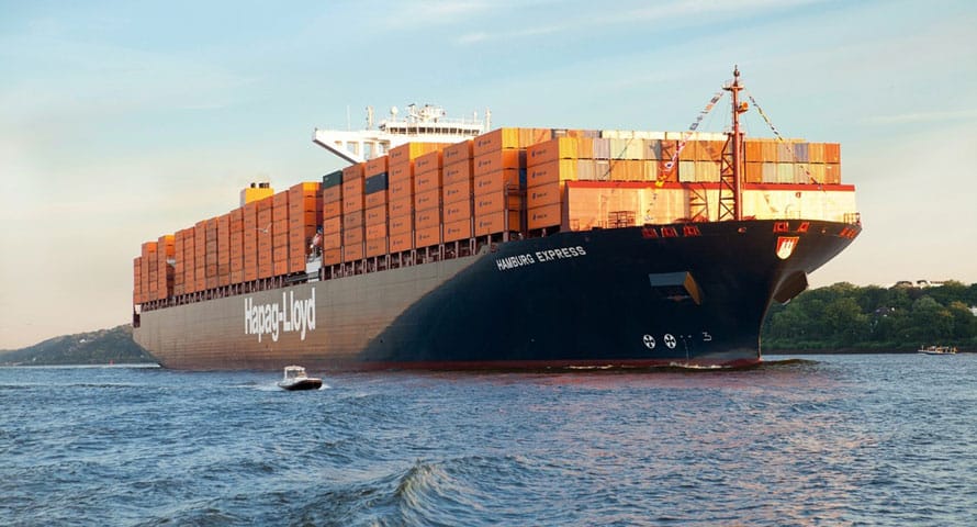A picture of a large black cargo vessel with neatly stacked containers on top being guided by a tugboat