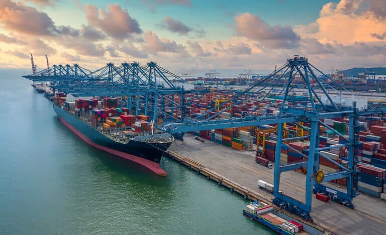 A large cargo vessel anchored at a harbour next to many containers in many colours
